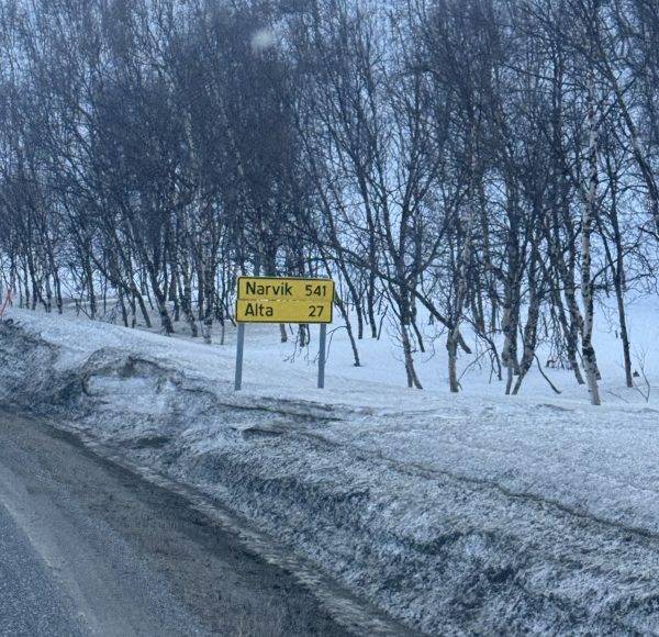 Straßenschild im Schnee neben einer winterlichen, mit kahlen Bäumen gesäumten Straße.