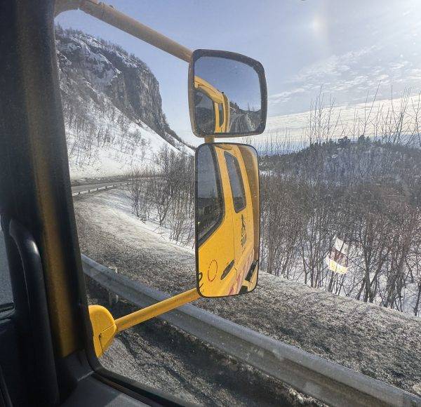Blick aus einem Fahrzeugfenster mit gelbem Seitenspiegel, verschneite Berglandschaft im Hintergrund.
