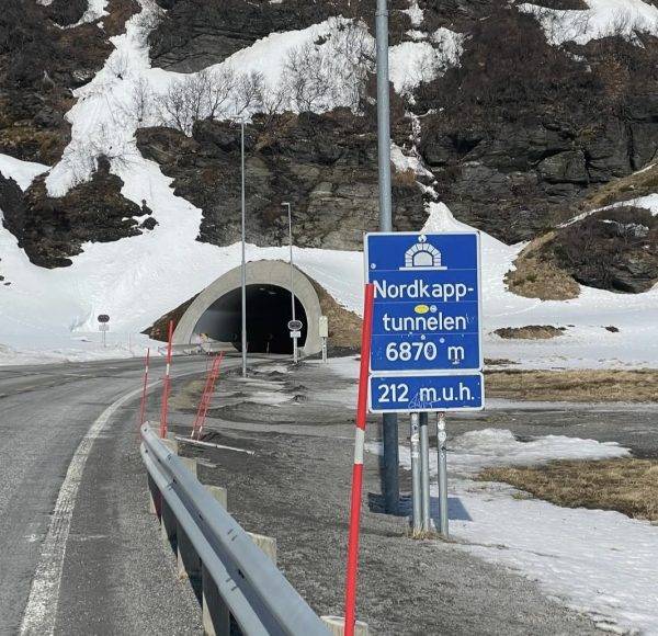 Straße führt zu einem Tunnel durch schneebedeckte Berge, blaues Verkehrsschild zeigt „Nordkapp-Tunnel 6870 m, 212 m u. M.“ an.