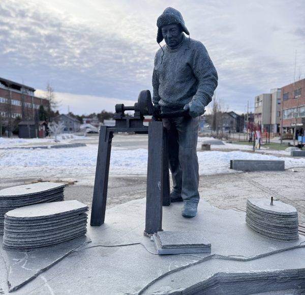 Statue eines Arbeiters mit Maschine, umgeben von Steinplatten auf einem Platz im Freien, Stadtgebäude im Hintergrund, winterliche Atmosphäre.