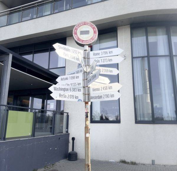Wegweiser mit mehreren Richtungsangaben vor einem modernen Gebäude. Ein Verkehrsschild ist oben auf dem Pfosten angebracht.