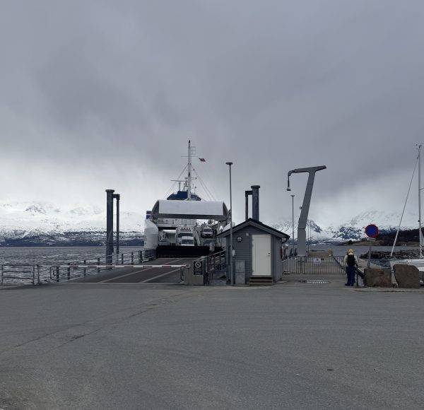 Eine Fähre liegt an einem grauen, bewölkten Tag am Dock, Berge sind im Hintergrund sichtbar.