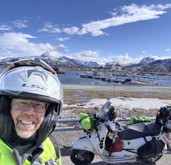 Motorradfahrer mit Helm macht Selfie vor schneebedeckten Bergen, See und geparktem Motorrad unter blauem Himmel.