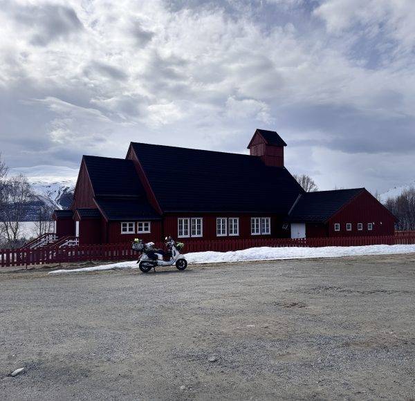 Rotes Holzgebäude mit dunklem Dach, Motorrad davor auf Parkplatz, Schnee am Rand, bewölkter Himmel im Hintergrund.