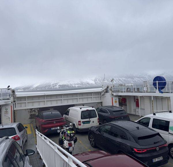 Mehrere Autos und ein Motorrad auf einer Autofähre bei bewölktem Himmel, im Hintergrund Schneelandschaft und Berge.