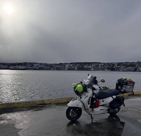 Motorrad mit Gepäck steht auf nasser Straße am Wasser, bewölkter Himmel, Stadt im Hintergrund.