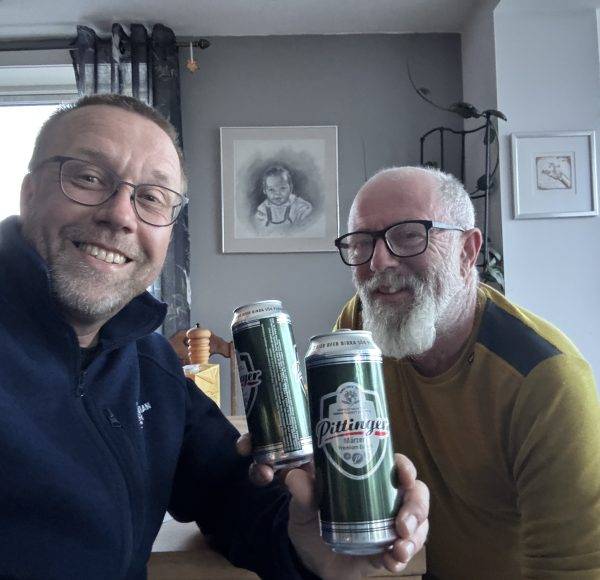 Zwei lächelnde Männer stoßen mit Bierdosen in einem Wohnzimmer an.