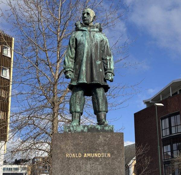 Bronzestatue eines Mannes in Winterkleidung steht auf einem Sockel mit der Aufschrift „Roald Amundsen“ in einer städtischen Umgebung.