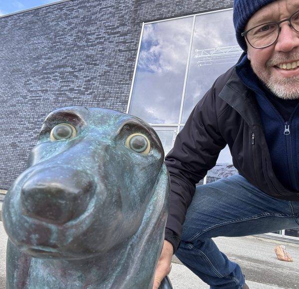Ein Mann mit Brille und Mütze posiert draußen neben einer Bärenstatue aus Metall.