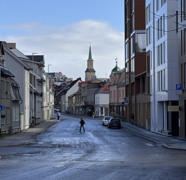 Leere Stadtstraße mit einzelnen Fußgängern, modernen und älteren Gebäuden, im Hintergrund eine Kirche mit Turm unter blauem Himmel.