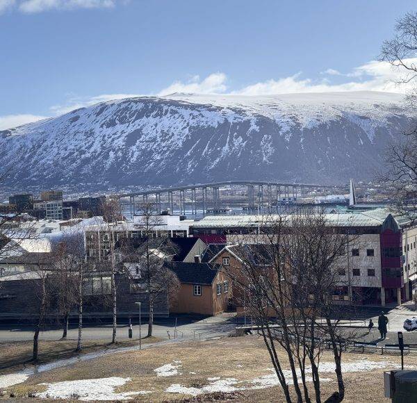 Stadt mit schneebedeckten Bergen im Hintergrund, einige Gebäude im Vordergrund, teilweise schneebedeckter Boden, blauer Himmel mit Wolken.