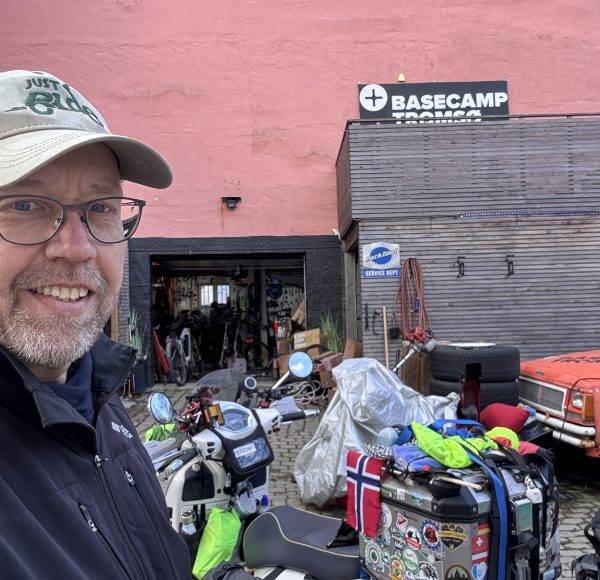 Mann mit Mütze lächelt vor einem Gebäude mit „Basecamp“-Schild und mehreren Motorrädern, die mit Gepäck beladen sind.