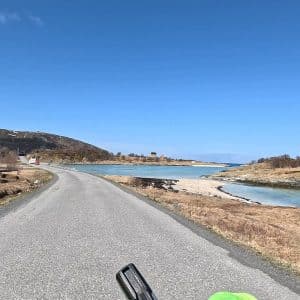 Asphaltstraße führt an Fluss und Hügeln entlang unter klarem, blauem Himmel.