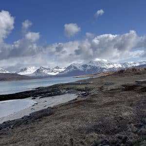 Schneebedeckte Berge, teilweise zugefrorener See, felsiges Ufer und wolkiger Himmel unter klarem Tageslicht.