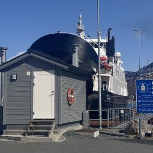 Ein graues Gebäude steht vor einem großen Schiff an einem sonnigen Tag, mit blauen Wegweisern und Bergen im Hintergrund.