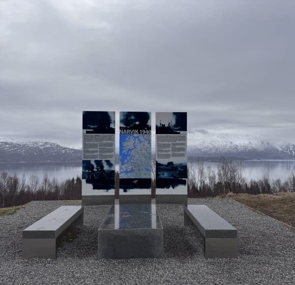 Spiegelnde Metalltafeln mit Informationstext und Bänken davor, auf einem Kiesplatz mit Bergen und einem See im Hintergrund unter bewölktem Himmel.