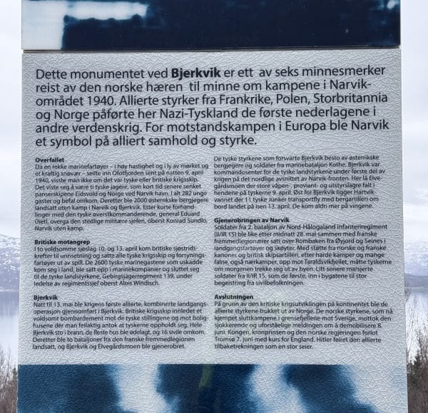 Informationstafel mit Text über das Kriegsdenkmal Bjørnevik in Norwegen, gewidmet der örtlichen Widerstandsbewegung im Zweiten Weltkrieg. Hintergrund leicht verschwommen, winterliche Landschaft.