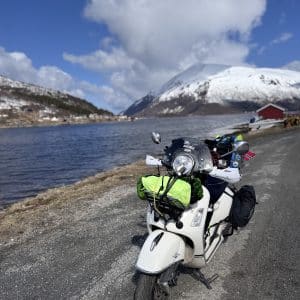 Motorrad mit Gepäck auf Straße neben See, im Hintergrund schneebedeckte Berge und bewölkter Himmel.