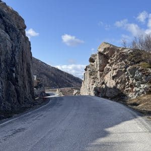 Straße führt durch felsige Schlucht, blauer Himmel mit wenigen Wolken, kahler Hang im Hintergrund.