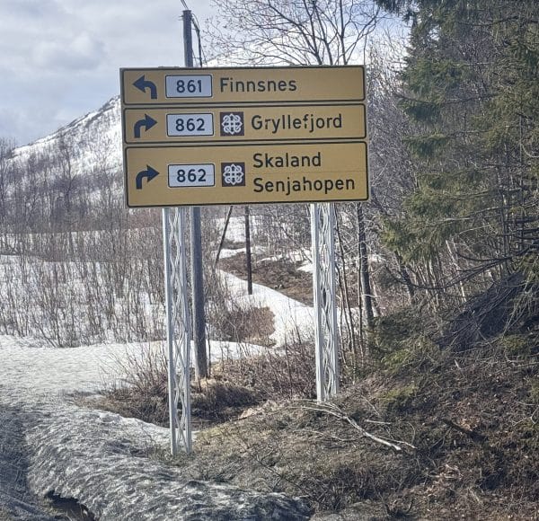 Straßenschild mit Richtungen zu Finnsnes, Gryllefjord und Sifjord, verschneite Landschaft und Bäume im Hintergrund.