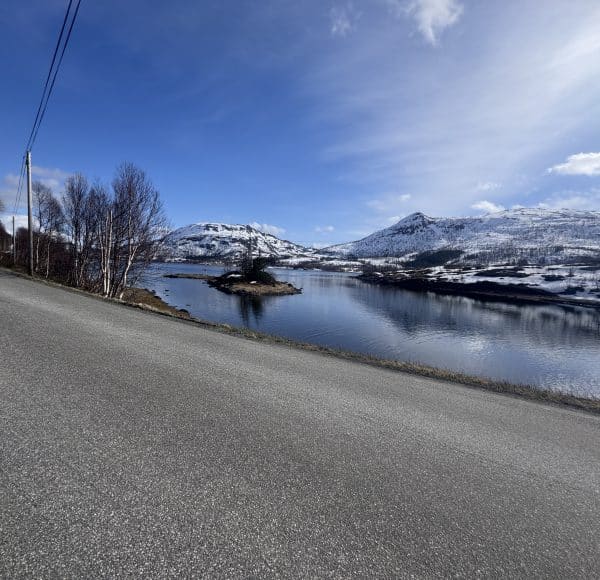 Straße am See mit schneebedeckten Bergen im Hintergrund und blauem Himmel.