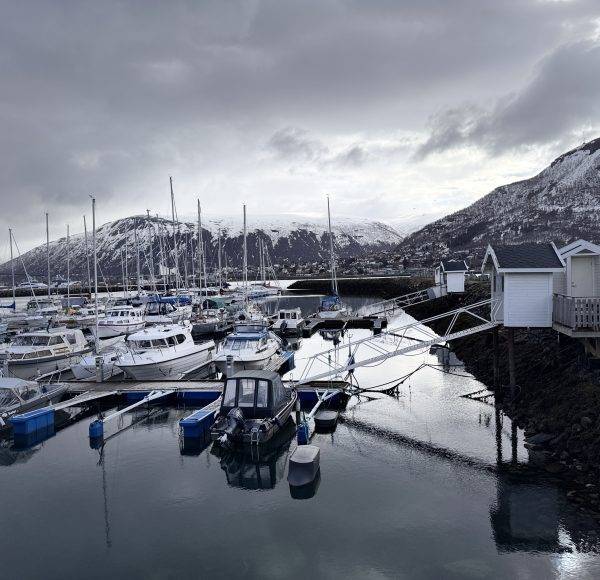 Boote liegen in einem ruhigen Hafen, umgeben von schneebedeckten Bergen unter bewölktem Himmel.