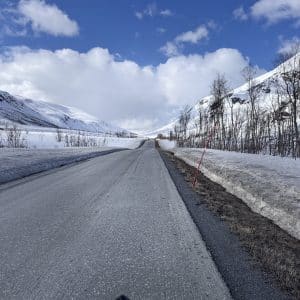 Verschneite Straße durch winterliche Berge, blauer Himmel mit Wolken, kahle Bäume am Straßenrand.