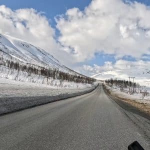 Eine gerade Straße führt durch eine verschneite Berglandschaft unter blauem Himmel mit weißen Wolken.