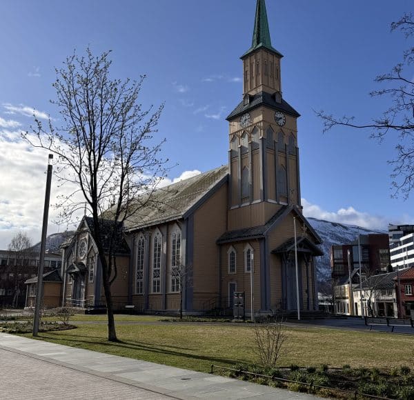 Kirche mit hohem Turm, umgeben von Bäumen und Wiese, im Vordergrund Gehweg, im Hintergrund Berge und moderne Gebäude.
