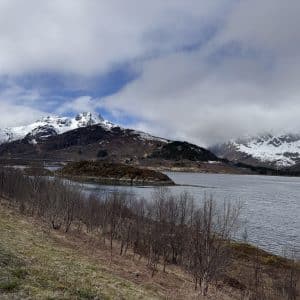 Schneebedeckte Berge im Hintergrund, ein See im Vordergrund, teils bewölkter Himmel, kahle Büsche entlang des Ufers.