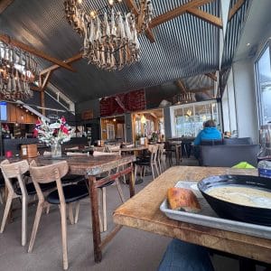 Gemütliches Café mit Holztischen, Kronleuchtern, großen Fenstern und einer Suppenschale auf einem Tablett im Vordergrund.