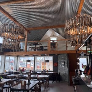 Modernes Restaurant mit hohen Decken, großen Fenstern, Holzdekor und zwei beleuchteten Kronleuchtern, Tische ordentlich angeordnet, gemütliche Atmosphäre.