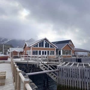Holzhaus am Wasser mit Veranda, umgeben von Zäunen und Stegen, bewölkter Himmel, Berge im Hintergrund.