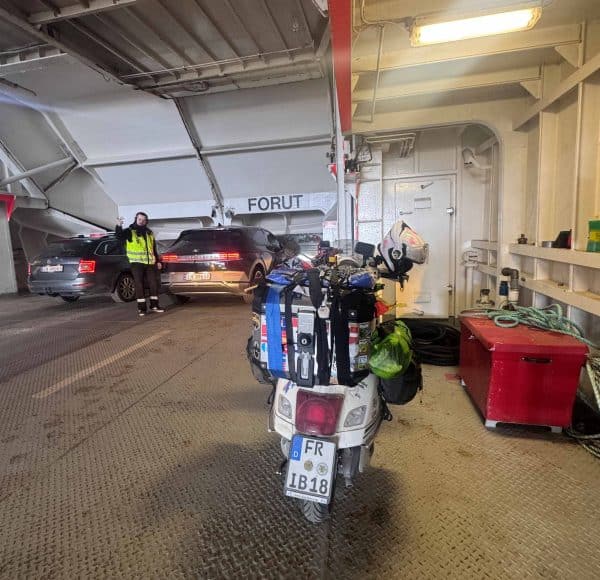 Motorrad mit Gepäck steht auf einer Autofähre, im Hintergrund parken Autos und ein Arbeiter in Warnweste ist zu sehen.