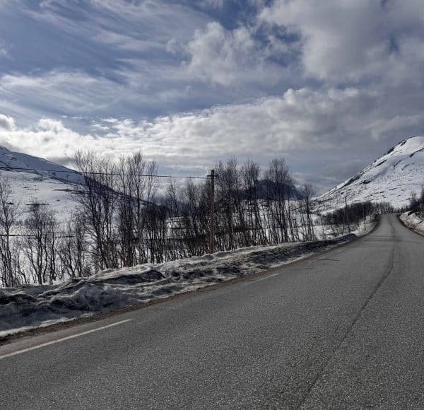 Leere Straße führt durch verschneite Berge mit kahlen Bäumen, teils bewölkter Himmel.