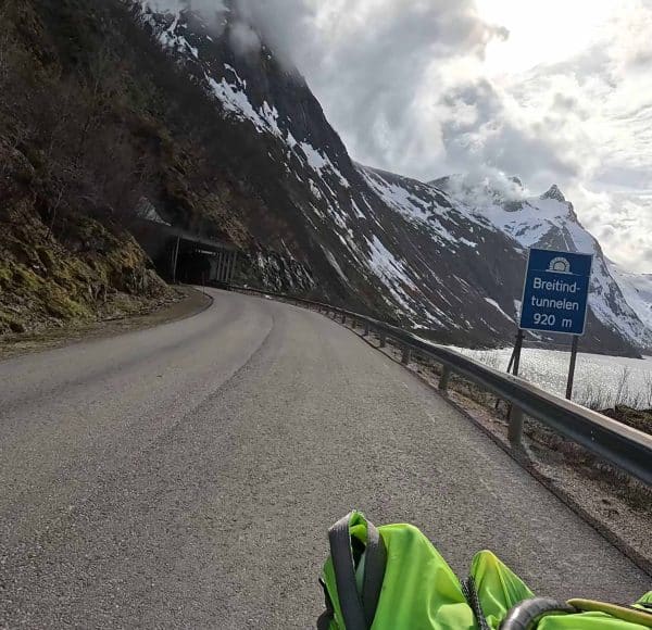 Straße führt durch bergige Landschaft mit Schnee, Tunnel voraus, Verkehrsschild auf Deutsch, Kurve nach links, Person mit leuchtender Jacke im Vordergrund, bewölkter Himmel.