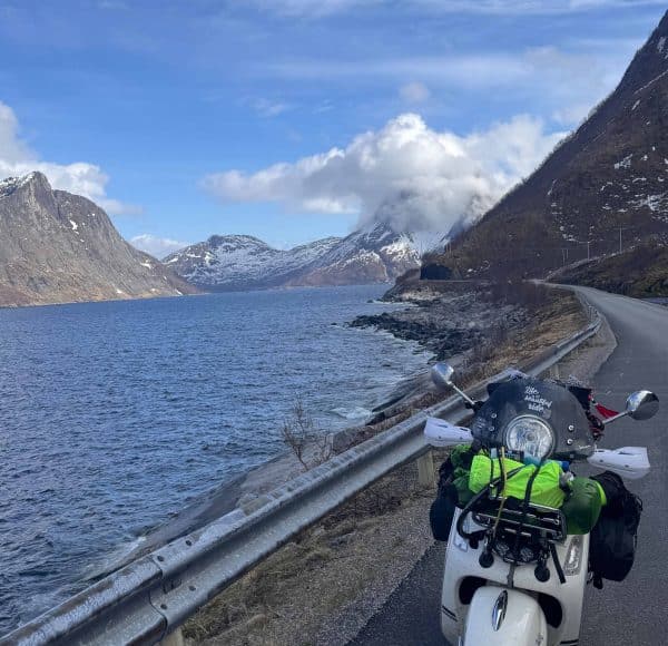 Motorradfahrer fährt auf einer Straße am See entlang, umgeben von Bergen und teilweise bewölktem Himmel.