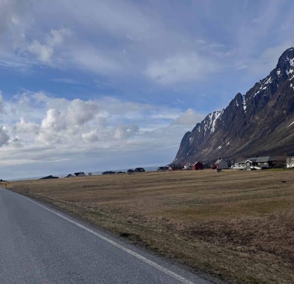 Straße mit Berglandschaft, einige Häuser am Fuß der steilen, schneebedeckten Berge unter bewölktem Himmel.