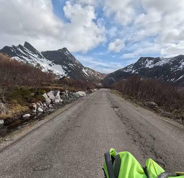 Straße führt durch bergige Landschaft mit schneebedeckten Gipfeln, blauer Himmel mit Wolken, vorne Teil einer grünen Jacke sichtbar.