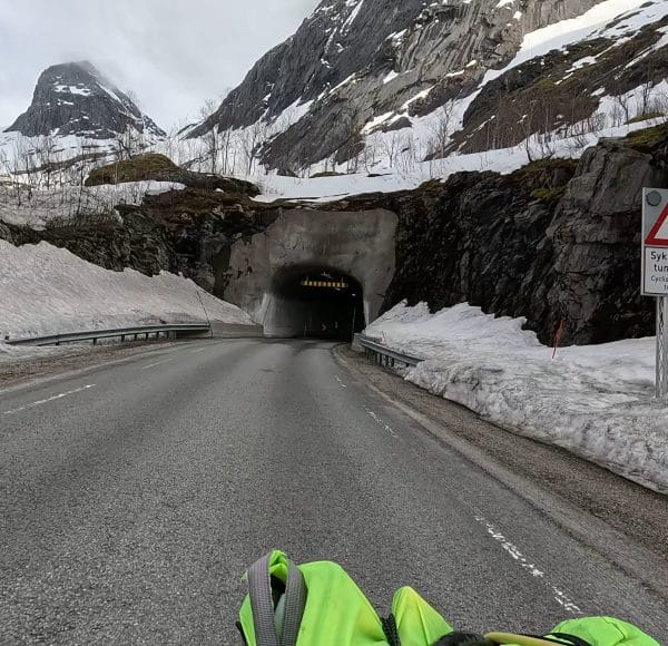 Straße führt zu einem Tunnel durch verschneite Berge, mit Schnee am Straßenrand und bewölktem Himmel, ein Motorradhelm ist im Vordergrund sichtbar.