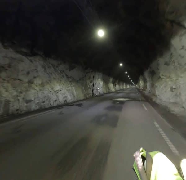 Straße führt durch einen beleuchteten Felstunnel, Wände aus rohem Stein, wenige Fahrzeuge sichtbar.