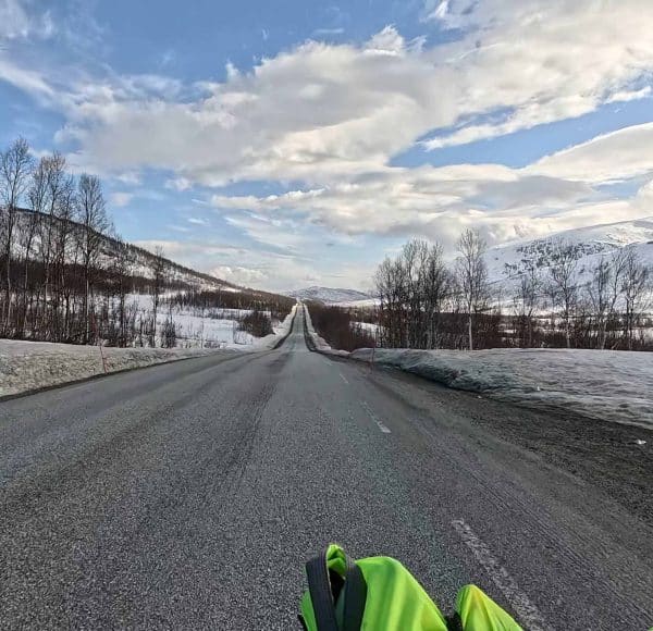Gerade Landstraße im Winter mit schneebedeckten Feldern und Bäumen, blauer Himmel mit Wolken, unten ist ein Teil einer grünen Jacke sichtbar.