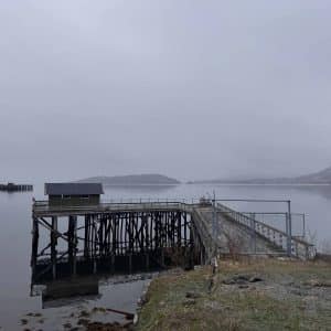 Ein hölzerner Steg führt auf einen nebligen See hinaus, im Hintergrund sind Berge im Nebel zu sehen.