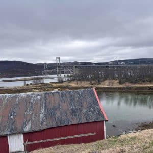Altes rotes Scheunengebäude am Wasser, hügelige Landschaft im Hintergrund, bewölkter Himmel.
