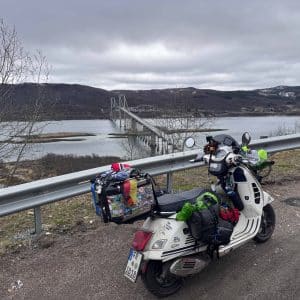Motorroller mit Gepäck steht an einer Straße vor einem Fluss und einer Brücke, umgeben von bewölktem Himmel und Hügeln.