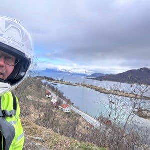 Motorradfahrer mit Helm vor einer malerischen Landschaft mit Wasser, Bergen und bewölktem Himmel.