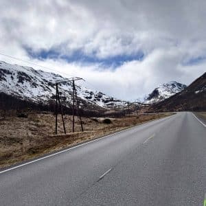 Leere Straße führt durch eine Berglandschaft mit schneebedeckten Gipfeln unter bewölktem Himmel.