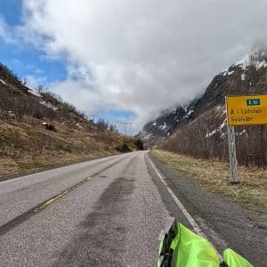 Leere Straße durch bergige Landschaft, bewölkter Himmel, Straßenschild auf der rechten Seite, teilweise schneebedeckte Berge im Hintergrund.