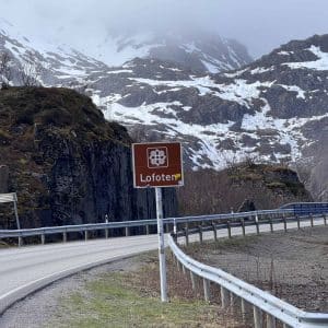 Straße mit Leitplanke, Verkehrszeichen „Lofoten“, dahinter verschneite Berge und bewölkter Himmel.