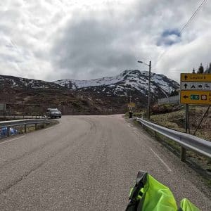Straße führt zu verschneiten Bergen, Verkehrsschilder am Straßenrand, bewölkter Himmel, fahrendes Fahrrad im Vordergrund.